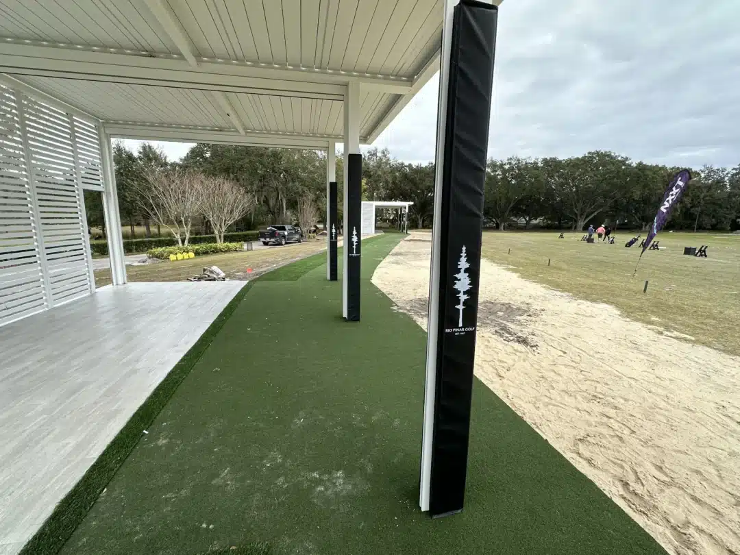 Covered outdoor golf practice area with white-painted beams, green artificial turf, and sandy terrain beyond; distant trees and players near a purple banner indicating a golf event or range setting.
