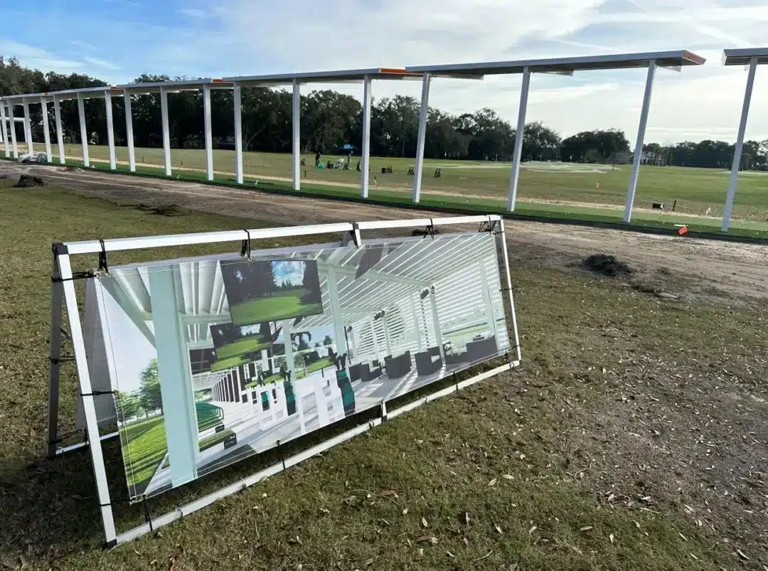 Row of white metal posts under construction in a park, with a large sign leaning on the ground showing interior architectural renderings.