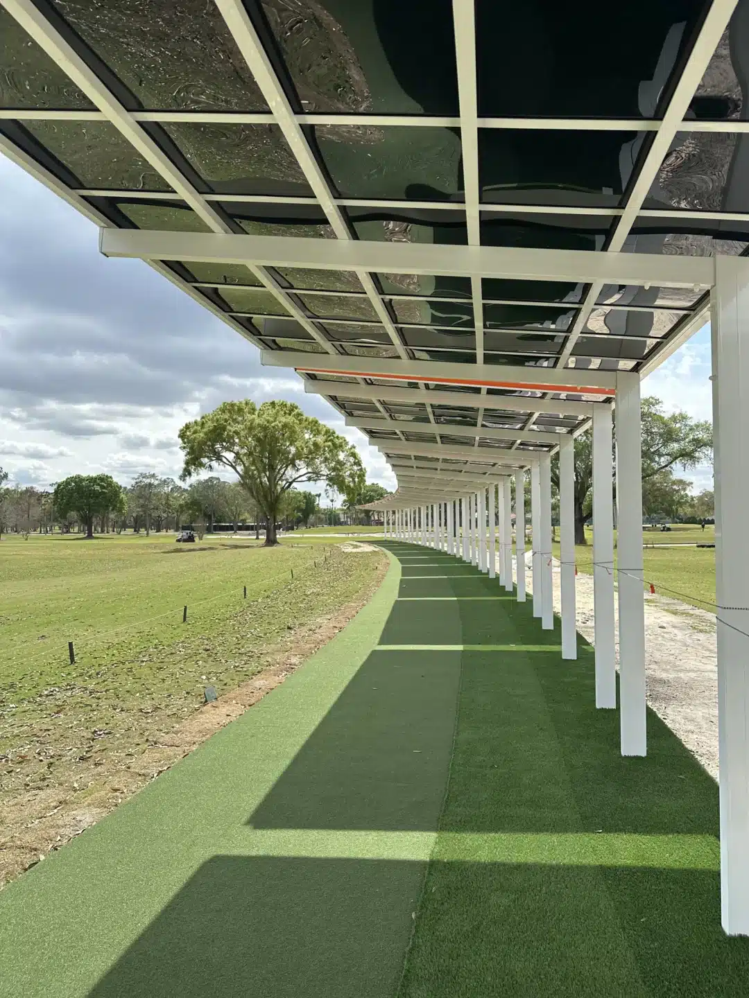 Long covered walkway with glass roof panels and white columns over a green artificial-turf path; trees in the distance.