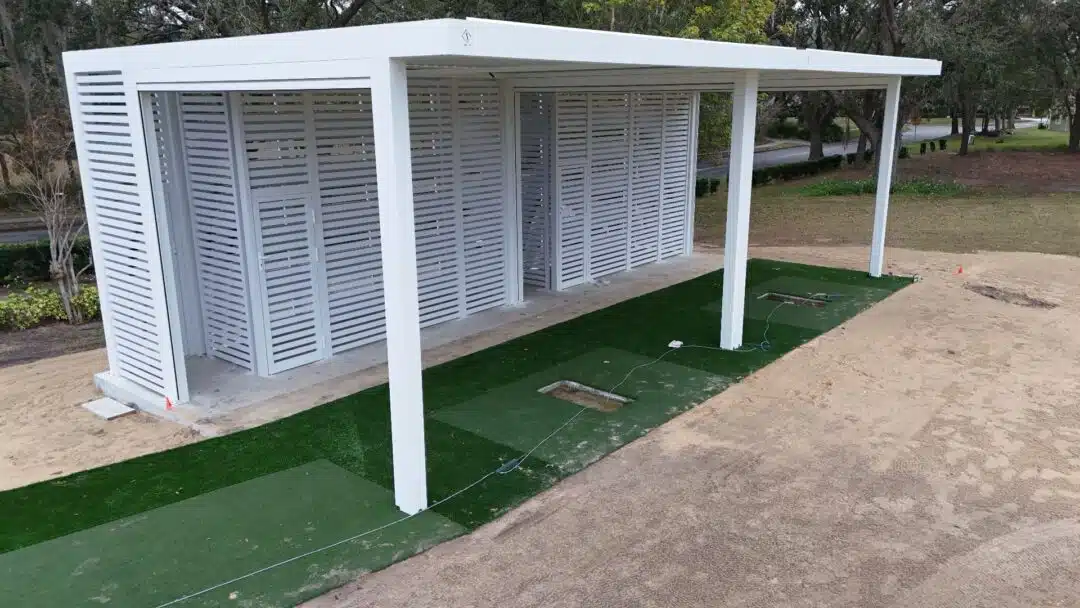White outdoor shelter with slatted, louvered walls and multiple bays for golf practice on green artificial turf mats in a sandy area