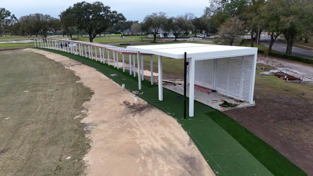 White, modern pavilion with slatted walls beside a curved, green-turf walkway in a park under construction.