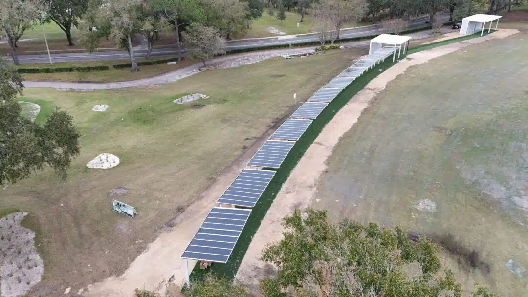 Aerial view of a long row of solar panels along a green walkway beside a sandy beach with trees nearby.
