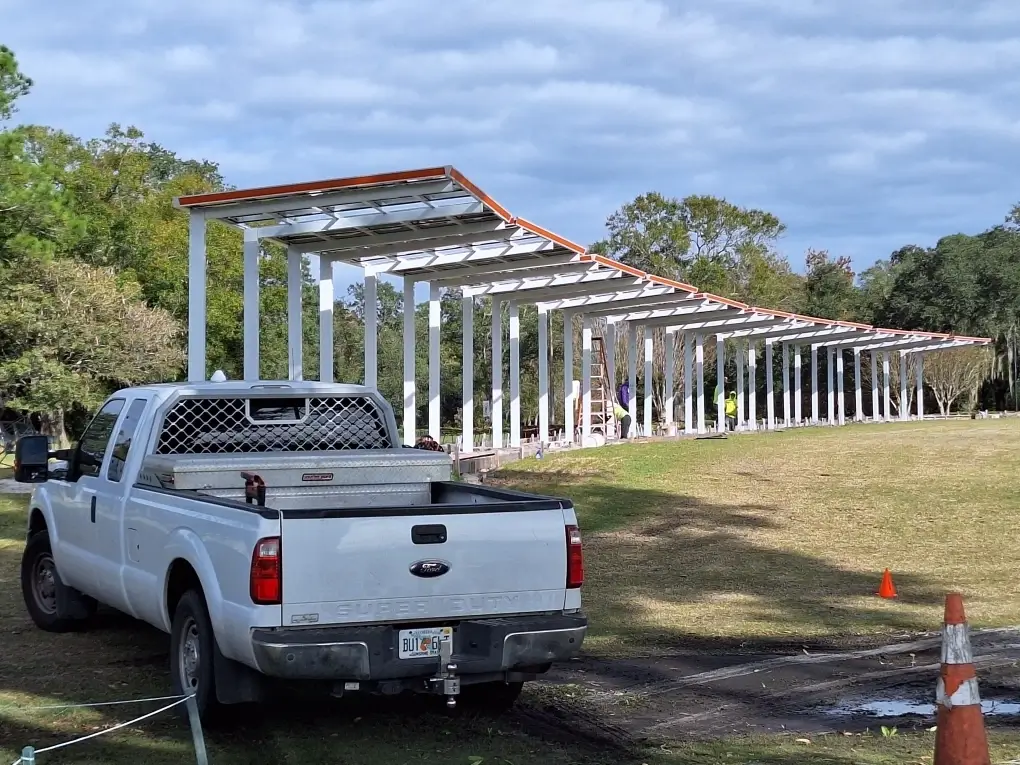White pickup truck parked on a grassy field beside a partially completed open-air metal shelter under construction; workers and ladder in the distance.