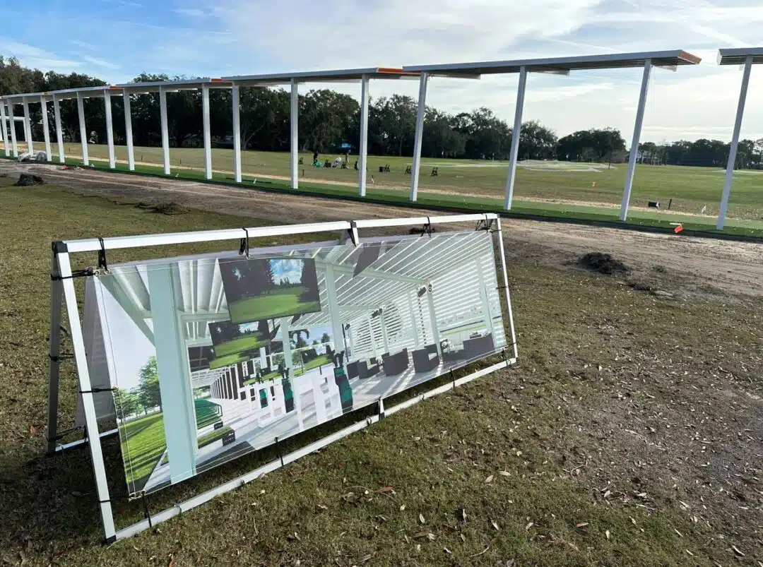 Row of white metal columns under construction in a grassy field, with a large framed render board leaning on a metal stand in the foreground.