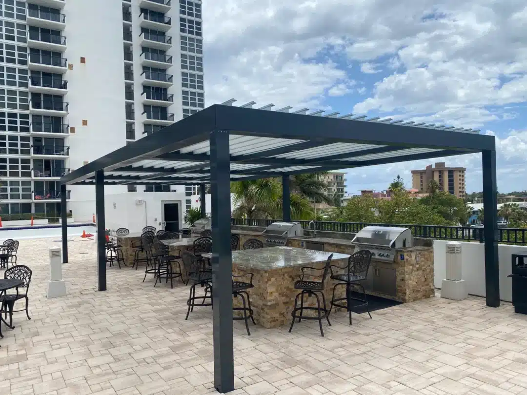 Outdoor communal grilling and seating area under a dark metal pergola, with built-in stone grills and wrought-iron chairs, high-rise buildings in the background.