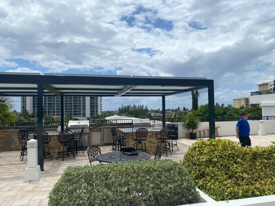 Outdoor patio with black metal chairs and round tables under a dark blue pergola; hedges in the foreground and buildings in the background under a cloudy sky.