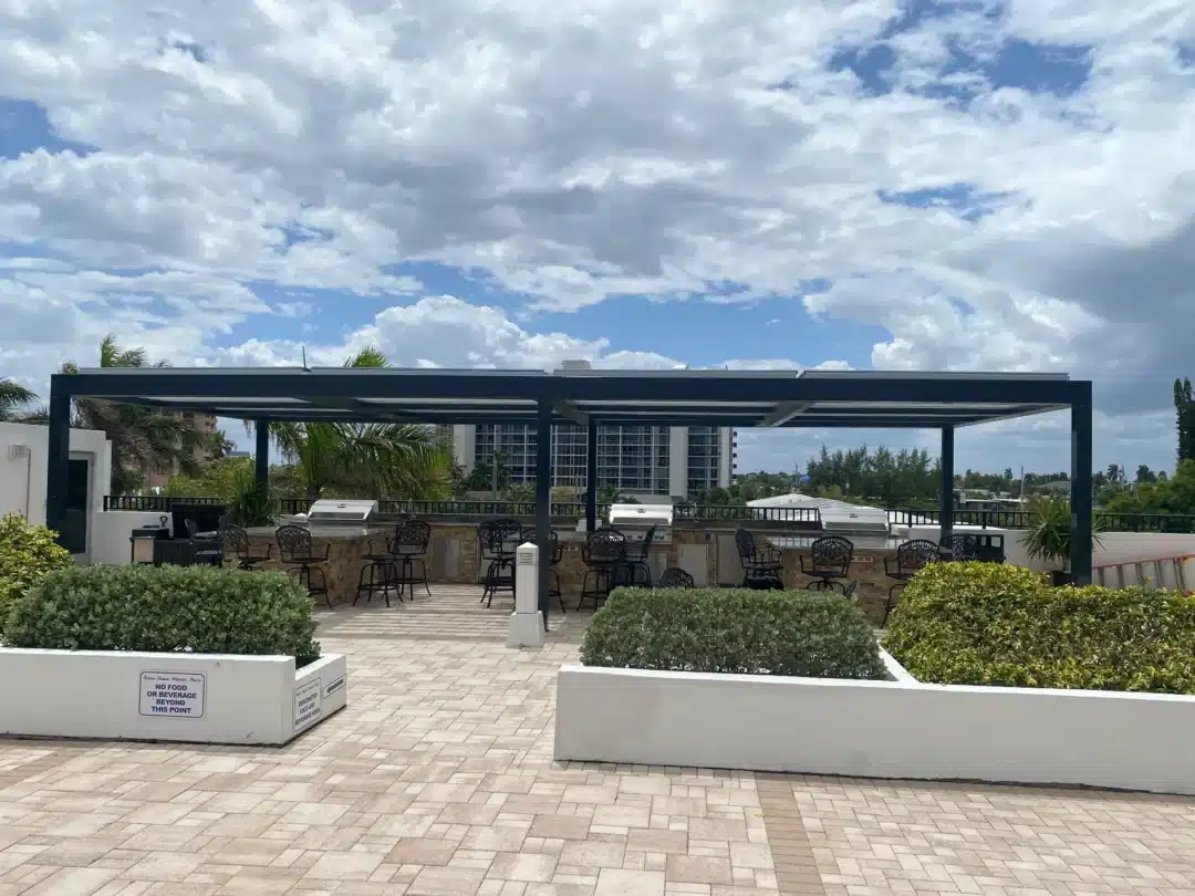 Outdoor bar and seating area under a dark metal pergola, with grills, tables, and hedges under a cloudy sky.