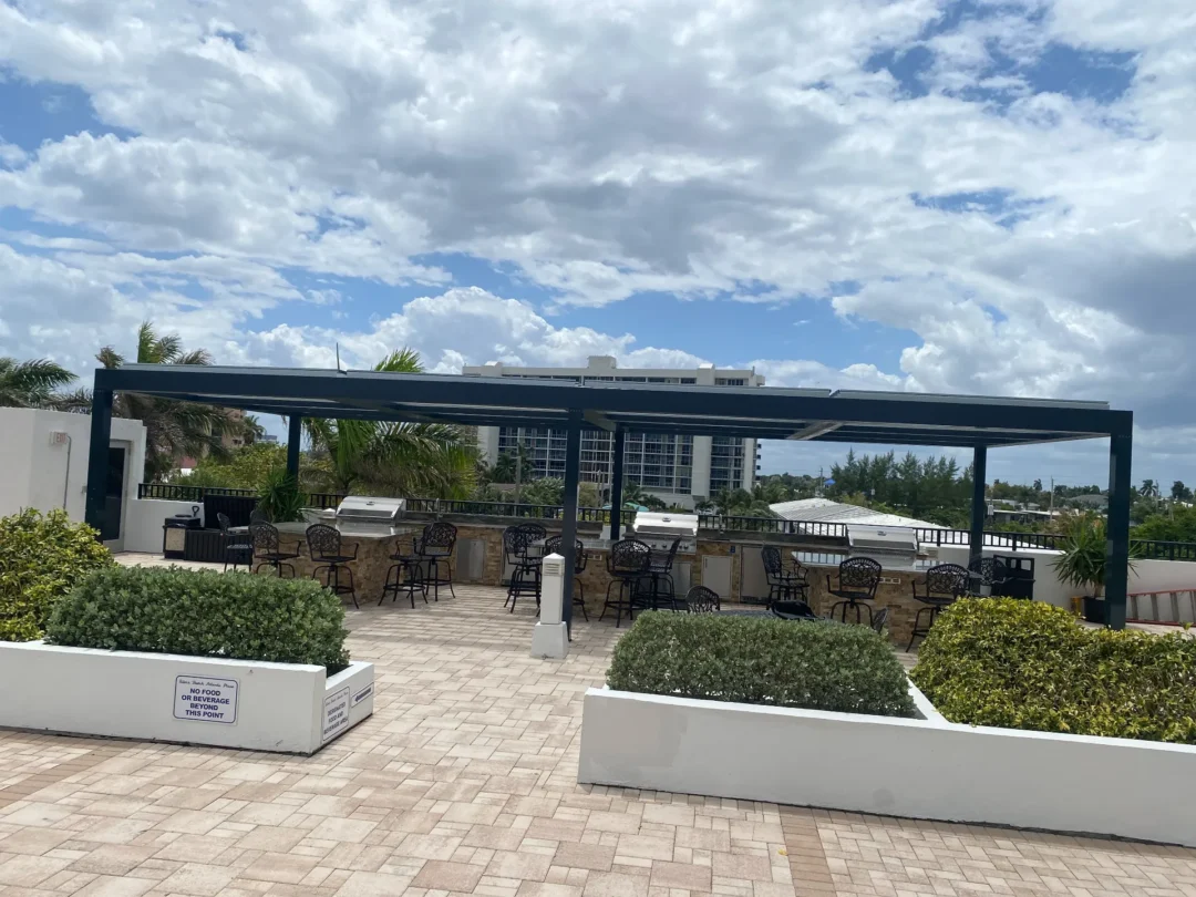 Outdoor patio area with built-in grills and numerous metal chairs under a dark blue pergola, palm trees, and a partly cloudy sky in the background.