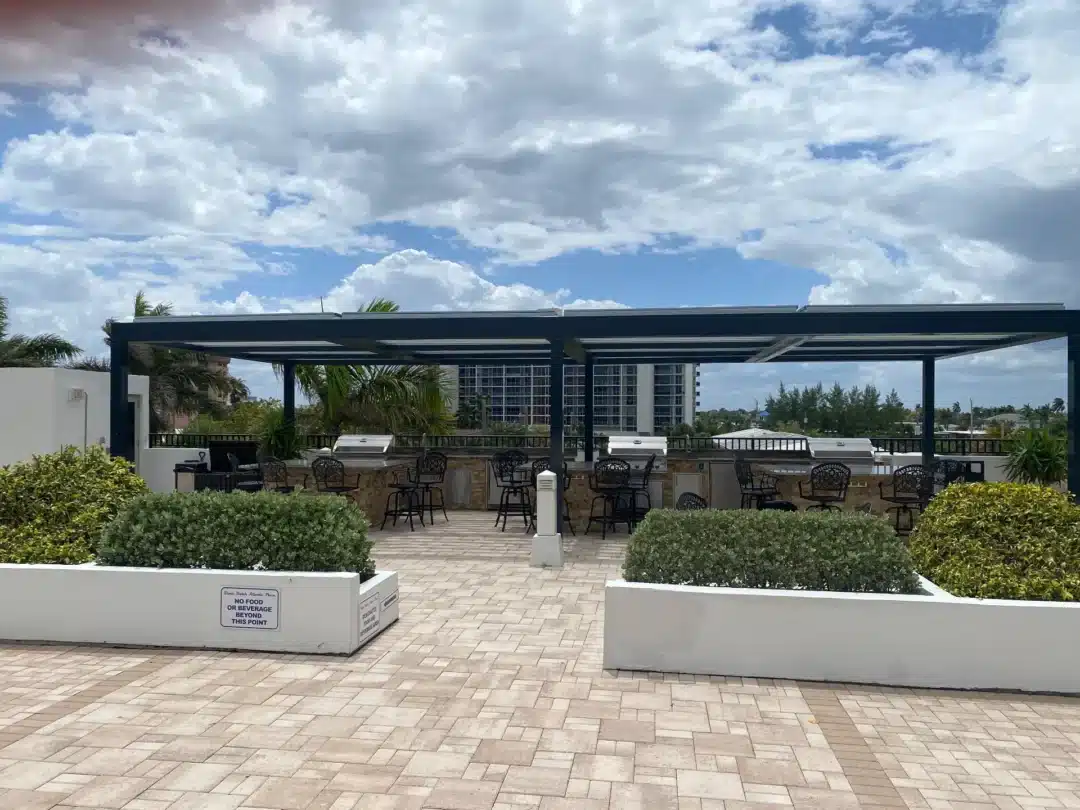 Outdoor bar or grill patio with black metal chairs under a blue pergola against a cloudy sky.