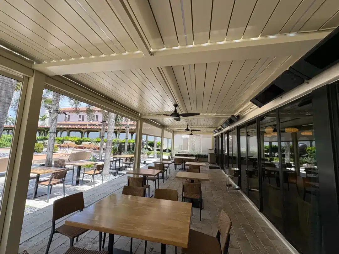 Outdoor cafe seating under a covered patio with wooden tables and wicker chairs, ceiling fans, and glass walls overlooking a sunny courtyard with palm trees.
