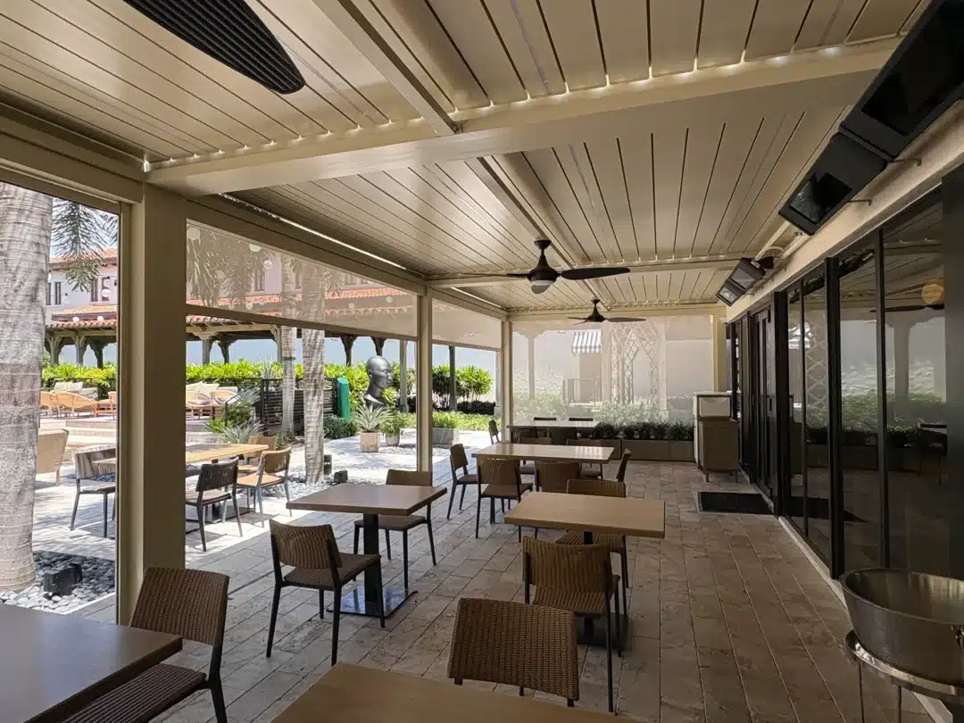 Outdoor cafe seating under a covered patio with wicker chairs and wooden tables, glass doors on the right, and palm trees outside
