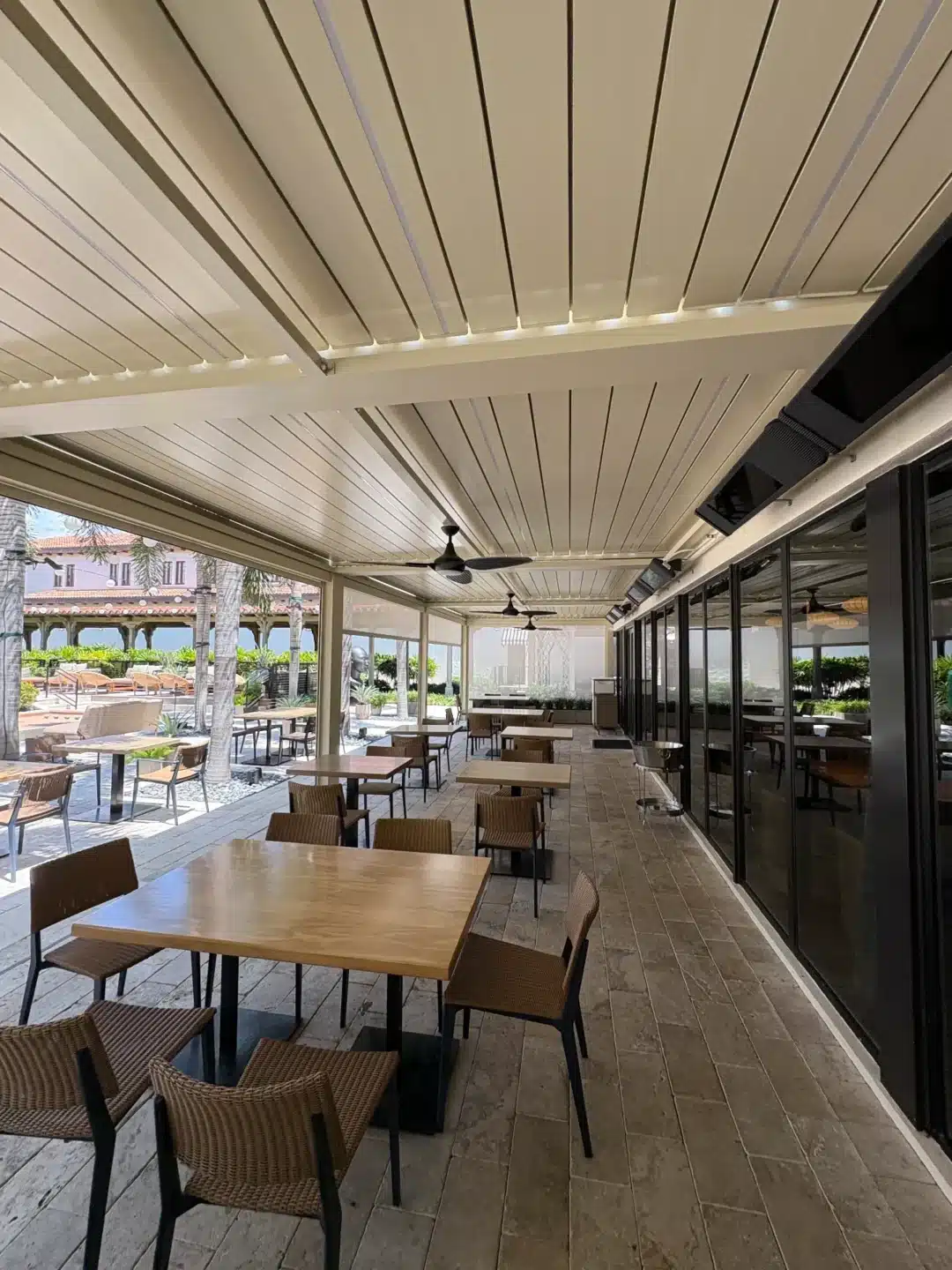 Outdoor dining area under a beige slatted canopy with wooden tables, wicker chairs, and ceiling fans beside glass-walled rooms.