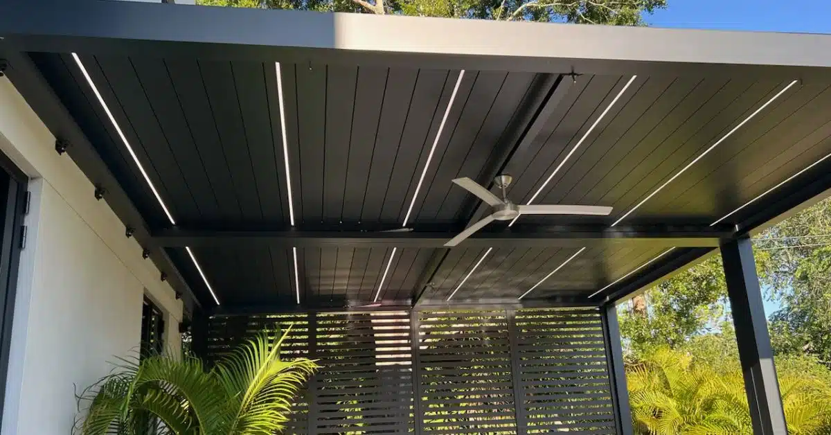 Modern Minimalist Pergola Lighting and Fan Integration - enVision Pergola A close-up of a modern black louvered pergola ceiling with integrated white linear LED lighting strips and a sleek silver ceiling fan.