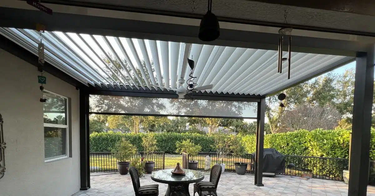 Comprehensive Outdoor Dining Pergola with Climate Control - enVision Pergola A wide view of a large dark-framed louvered pergola installed over a stone patio dining area, featuring an integrated white ceiling fan and wind chimes.